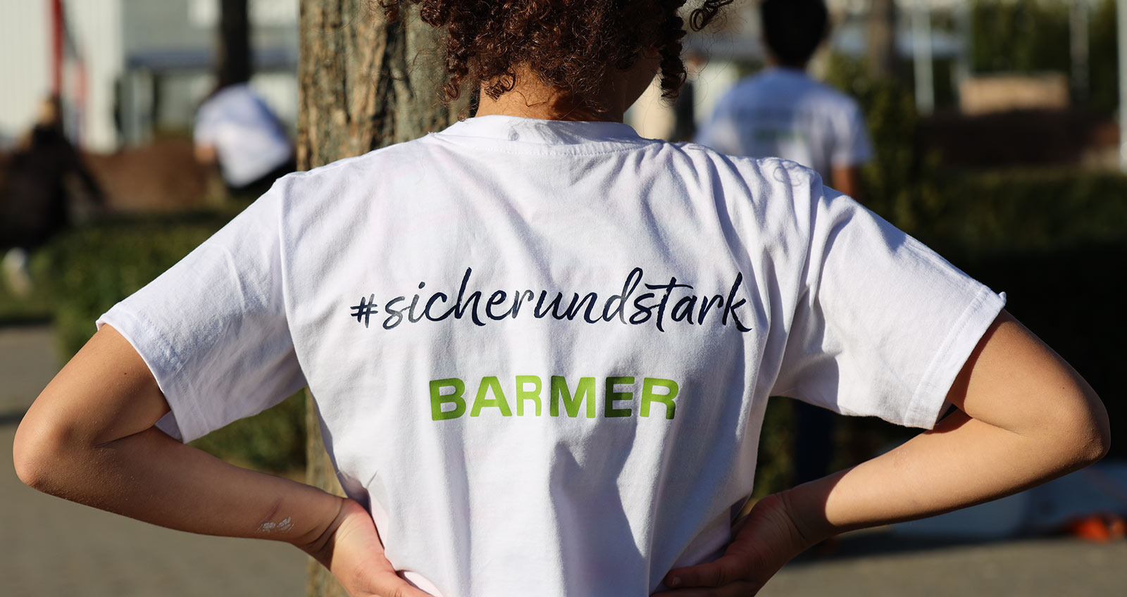 Ein Kind in einem weißen T-Shirt ist von hinten zu sehen, auf dem Shirt steht: "#sicherundstark", dadrunter das Logo der "Barmer"