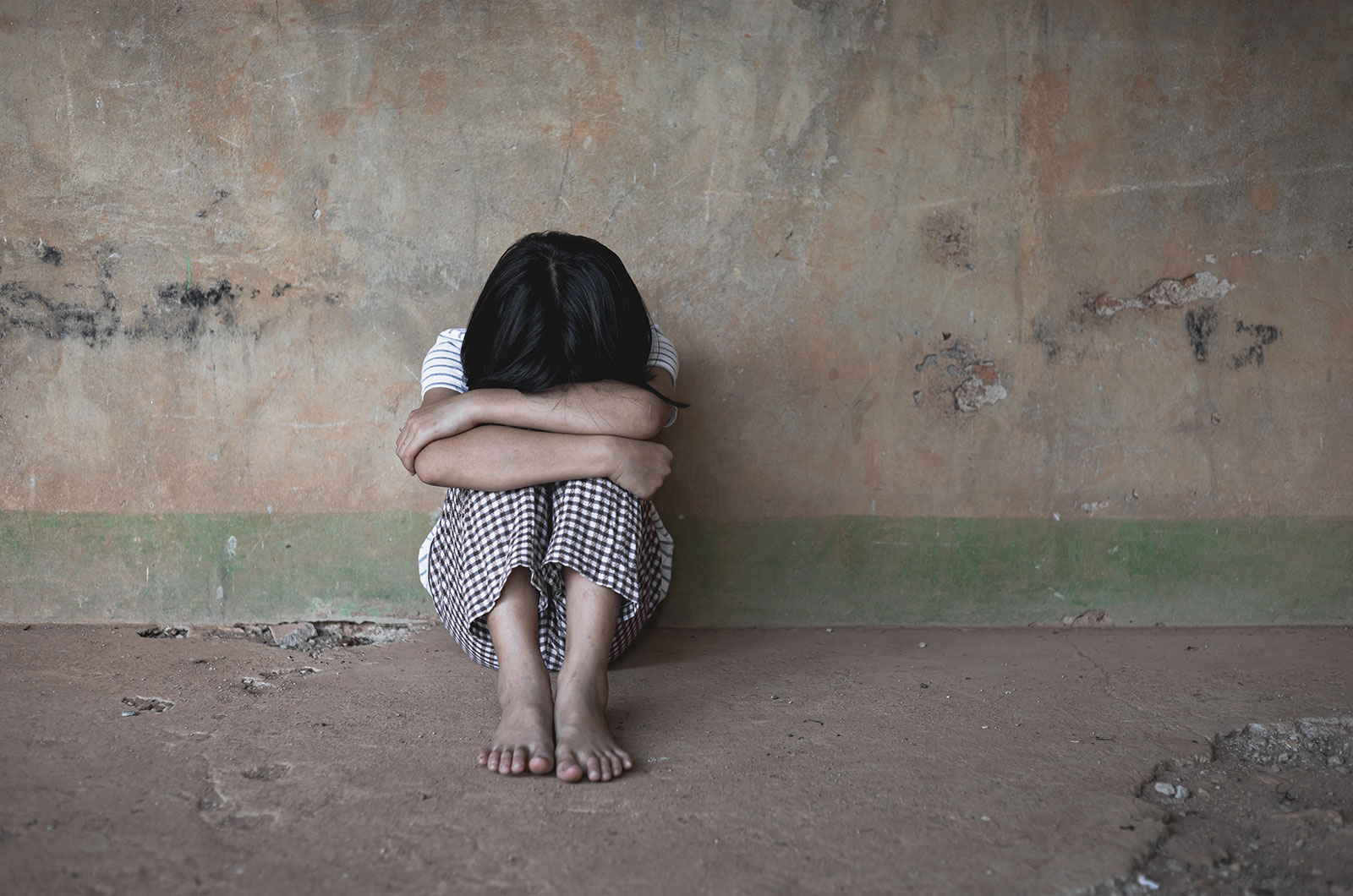 einsames, armes, obdachloses, verzweifeltes, traumatisiertes Kind, sitzt barfuß vor einer kargen, schmutzigen Wand, den Kopf auf die Knie gestützt, mit beiden Armen fest umschlungen