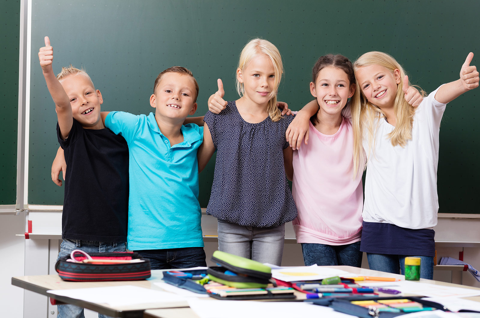 Gruppe fröhlicher Schulkinder vor Schultafel mit Daumen nach oben 