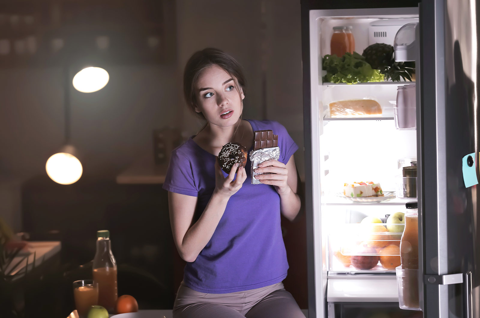 junge Frau, die nachts heimlich vor dem Kühlschrank ungesundes Essen, Schokolade und einen Donut, in der Küche isst