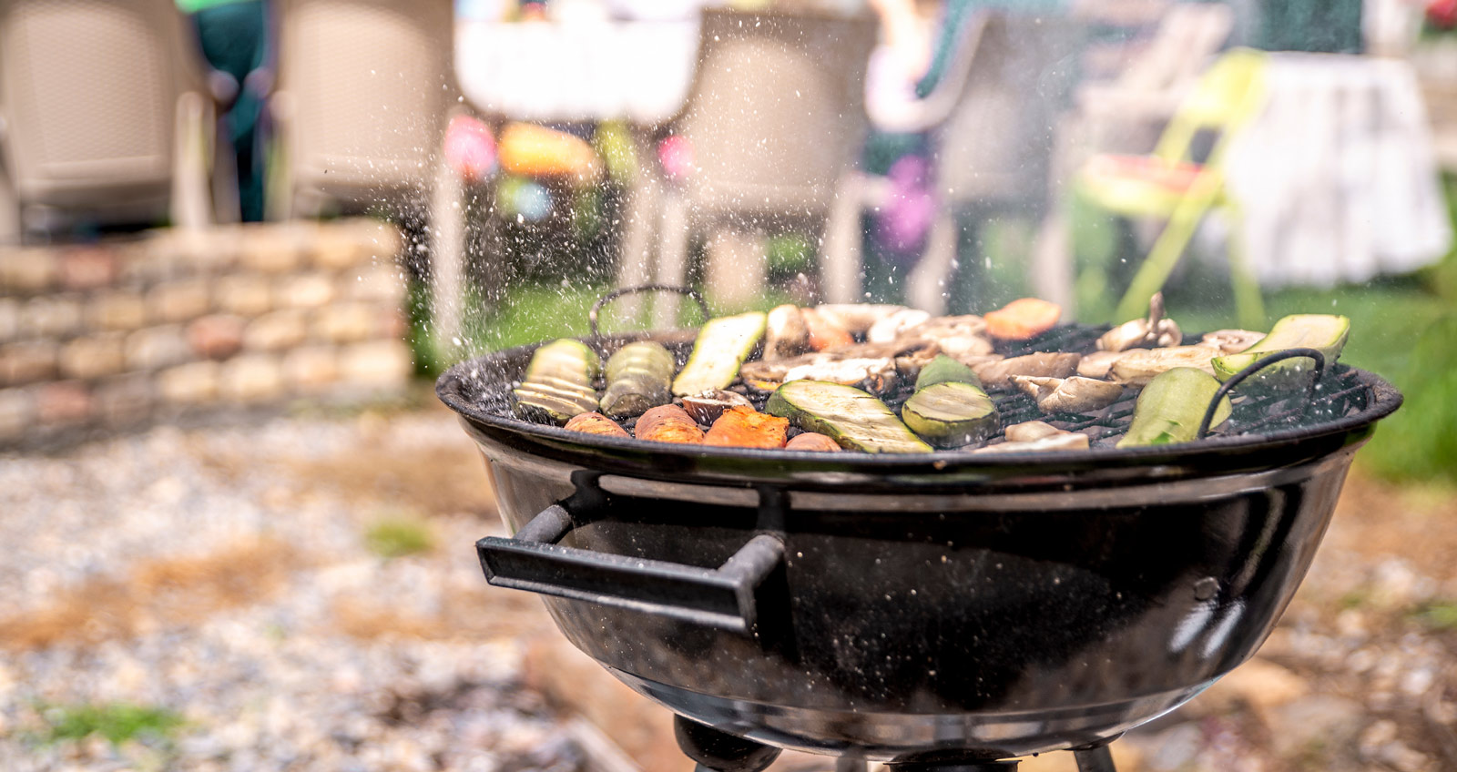 Ein Grill voller Gemüse und ein bisschen Fleisch vor einer Gruppe fröhlicher Menschen im Garten