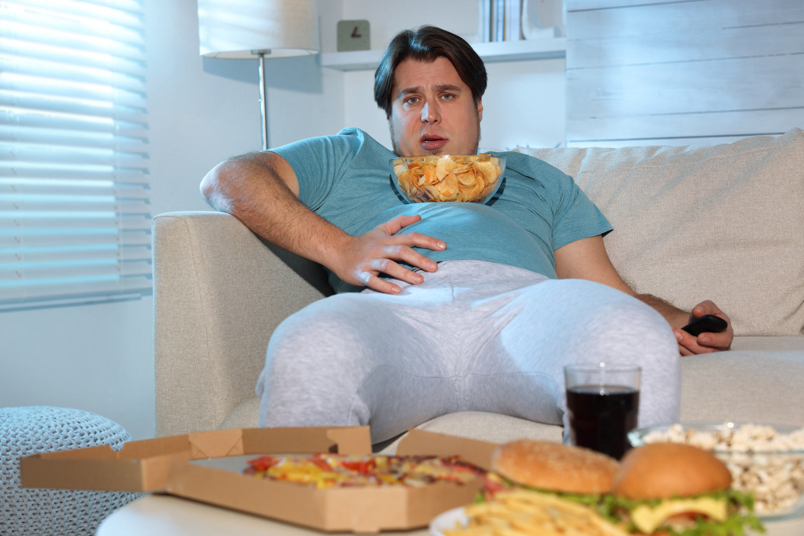 EIn Mann sitzt vollgefressen auf dem Sofa umgeben von Fast Food