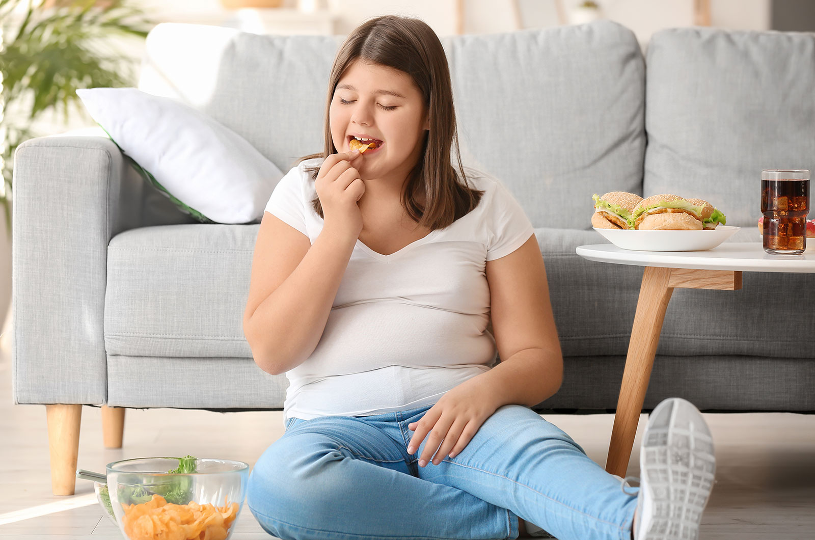 übergewichtiges Mädchen, sitzt vor der Couch und isst Chips 